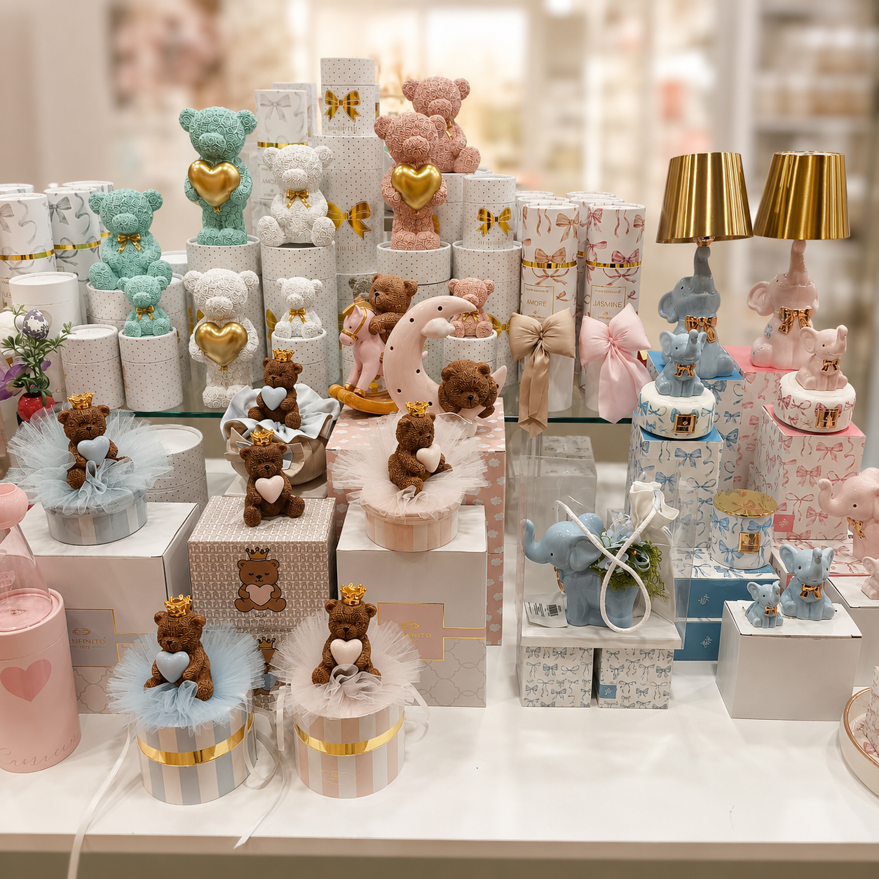 Esposizione di bomboniere e idee regalo con orsetti decorativi, elefantini, scatole regalo e accessori nei toni pastello rosa, azzurro e oro.
