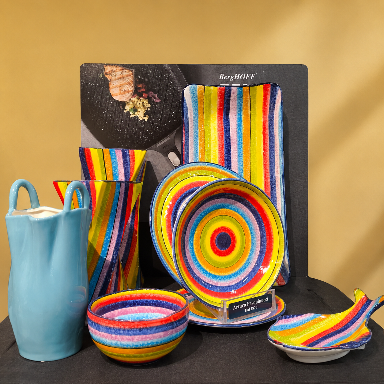 Set di piatti e accessori da tavola in ceramica multicolore artigianale con motivi arcobaleno, esposti su fondo beige.